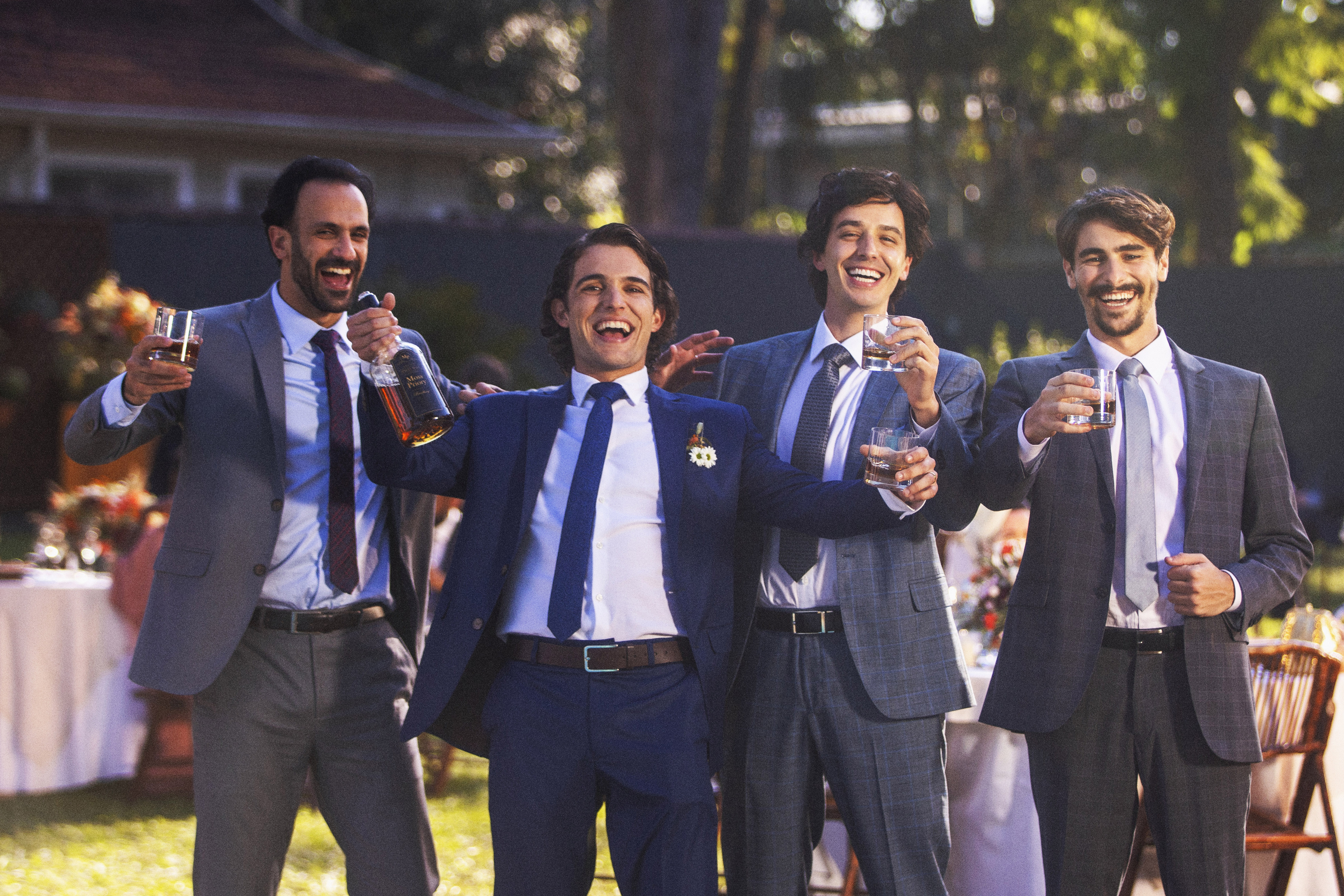 Quatro homens vestidos de terno posam para foto com bebidas na mão em uma festa de casamento